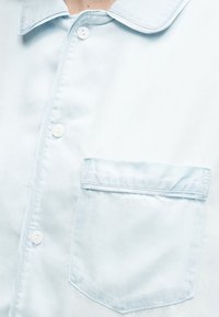 Κοντινό πλάνο ενός ανοιχτό μπλε πουκάμισου με μυτερό γιακά και μία τσέπη στο στήθος στη δεξιά πλευρά.