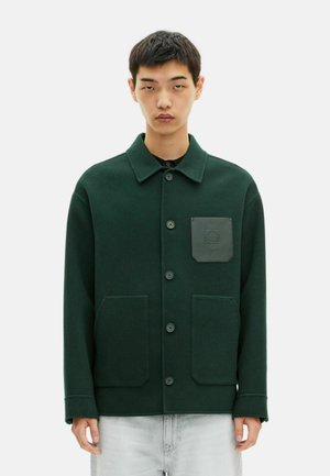 Junger Mann, der einen dunkelgrünen Hemdjacke mit einer schwarzen Emblem-Patch-Tasche und hellgrauen Jeans trägt, vor einem weißen Hintergrund steht.