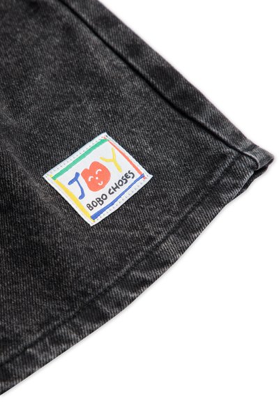 Harmaan denim-kangas, jossa on värikäs etiketti, jossa lukee "JOY" ja "Bobo Choses". Etiketti on hymyilevällä naamalla ja suorakulmaisella muodolla.