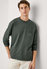 Camisa de manga larga verde oscura de algodón, con un bolsillo en el pecho que tiene una etiqueta, cuello acanalado y un corte relajado.