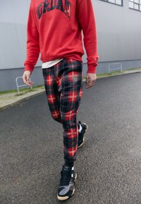 Rode sweatshirt met het "JORDAN" logo, gecombineerd met taps toelopende geruite broek in marineblauw, rood en zwart, en zwarte sport sneakers.