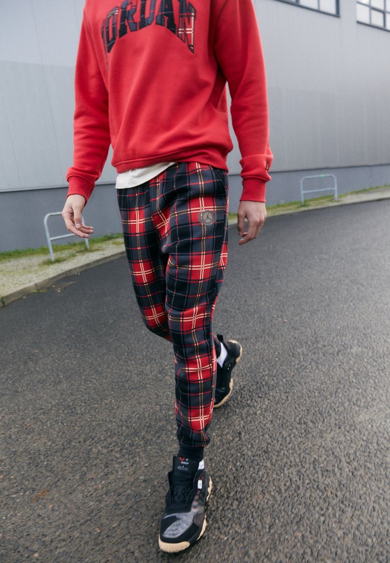 Rode sweatshirt met het "JORDAN" logo, gecombineerd met taps toelopende geruite broek in marineblauw, rood en zwart, en zwarte sport sneakers.