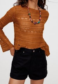 Maglione all'uncinetto a trama aperta color arancio bruciato con maniche a campana lunghe, indossato sopra pantaloni corti in denim neri con risvolti e chiusura a bottoni.