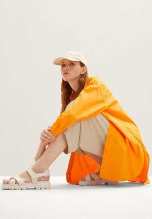 Bright orange oversized shirt, beige shorts, and cream sandals with thick soles. Model crouches on a neutral background.