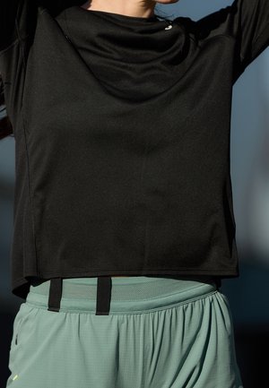 Zbliżenie osoby noszącej czarną bluzkę z długim rękawem i jasnozielone szorty sportowe, z rękami uniesionymi nad głową na rozmytym tle.