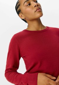 Pull en maille rouge avec un col rond et des manches longues. Il présente une texture lisse et des poignets côtelés. La coupe semble ajustée au corps.