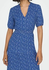 Sininen kukkamekko, jossa on lyhyet puffihihat, V-kaula-aukko ja istuva vyötärö. Kangas on koristeltu pienellä valkoisella lehtikuviolla kauttaaltaan.