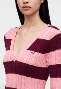 Jeune femme portant un pull en maille torsadée rayé rose et bordeaux avec un col en V, visible des épaules à la poitrine.