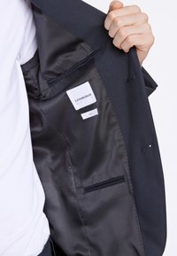 Blazer blu navy con fodera interna liscia; presenta una tasca sul petto e etichette. Il tessuto ha una trama con una luminosità discreta.