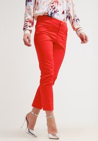 Pantaloni aderenti rossi con una texture liscia e due tasche laterali, abbinati a una blusa colorata e stampata e scarpe con tacco alto argentate.