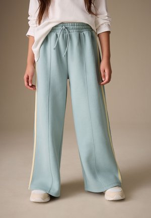 Person iført lyseblå vidde sweatpants med gule sidestriber, hvide sneakers og en hvid langærmet skjorte med ærmerne rullet op.