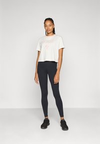 Tricou scurt de culoare crem cu logo-ul roz "GUESS", asociat cu colanți negri și pantofi sport negri, pe un model pe un fundal simplu.
