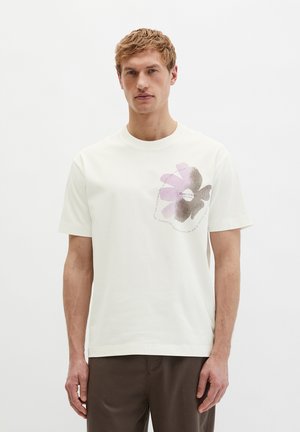 Blonder Mann trägt ein weißes T-Shirt mit einem großen pinken und grauen Blumenmuster auf der Brust und dunkelbraune Hosen, steht vor einem weißen Hintergrund.
