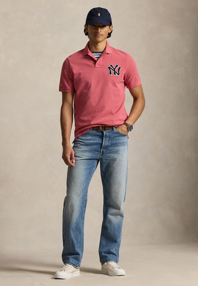 Άνδρας που φοράει ροζ πόλο με μαύρο λογότυπο NY, ανοιχτό μπλε τζιν, μπεζ αθλητικά παπούτσια, μπλε σκουφάκι και ρολόι χειρός, που στέκεται μπροστά σε φόντο μονόχρωμο.