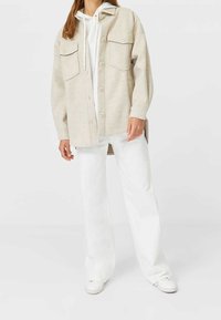 Lichtbeige oversized shirtjacket met twee voorzakken, knoopsluiting en een zachte textuur, gecombineerd met witte broek en schoenen.