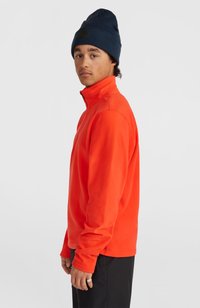 Oranje fleece pullover met een hoge kraag, voorzien van een ritssluiting aan de voorkant en lange mouwen. Draagt een marineblauwe beanie. Zwarte broek.