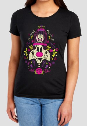 Frau trägt ein schwarzes Shirt mit buntem Cartoon-Hundegesicht, rosa Nase, Totenkopf in einem rosa Vogelkäfig darüber, umgeben von gelben und violetten Blumenmustern.