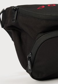 Μαύρο υφασμάτινο τσαντάκι μέσης με υφή στην επιφάνεια, διαθέτει θήκες με φερμουάρ και κόκκινη κεντητή λεπτομέρεια. Συμπεριλαμβάνεται ρυθμιζόμενος ιμάντας.