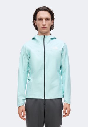 Mann mit hellblauer Kapuzenjacke mit Reißverschluss und dunkelgrauer Hose, steht vor einfachem hellem Hintergrund.