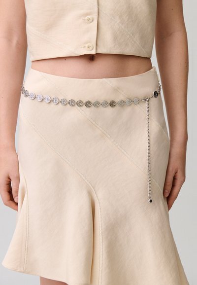 Completo beige a due pezzi con top corto abbottonato e gonna svasata, accessoriato con una cintura a catena argentata con monete intorno alla vita.