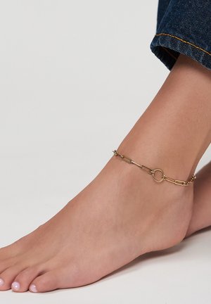 Barfuß mit einer goldenen Gliederfußkette mit rundem Verschluss, vor einem schlichten weißen Hintergrund und dunkelblauen Jeans.