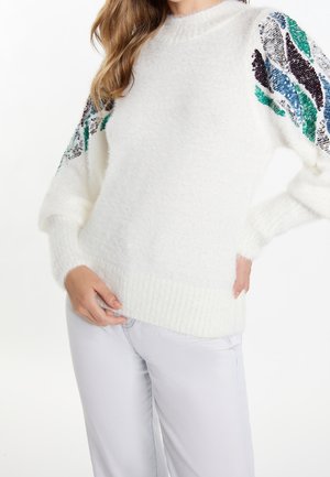 Pull blanc duveteux avec motifs multicolores en sequins sur les manches bouffantes, porté avec un pantalon gris clair, mains reposant près de la taille.