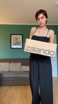 Krabica Zalando z lepenky, ktorú drží osoba, svetlohnedá farba s tučnými sivými písmenami. Pozadie zobrazuje zelenú stenu a sivú pohovku.