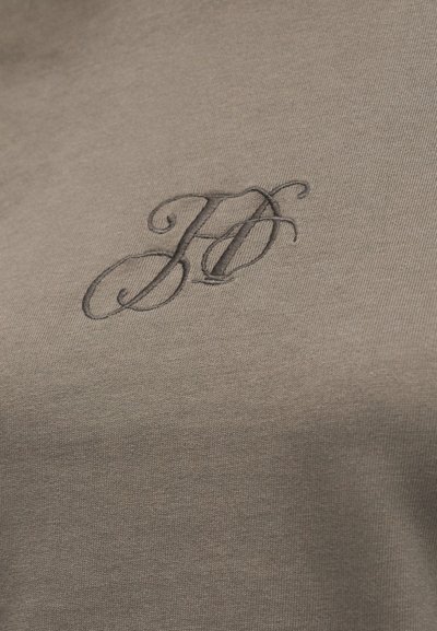 Κοντινή λήψη υφάσματος ταφτής με σκουρόχρωμα κεντημένα αρχικά «K» και «F» σε καλλιγραφική γραφή στην περιοχή του στήθους.