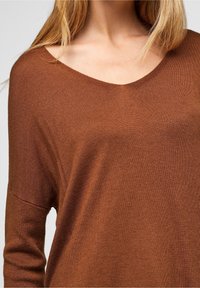 Pull en maille marron douce avec un large col en V et des manches amples. Le tissu présente une texture subtile et une coupe décontractée.