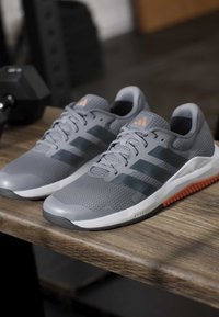 Ζευγάρι γκρι παπούτσια Adidas για τρέξιμο με μαύρες ρίγες και πορτοκαλί λεπτομέρειες τοποθετημένα σε ξύλινο πάγκο σε γυμναστήριο.