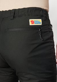 Čierne nohavice Fjällräven s nášivkou, elastickým pásom a zipsovým vreckom na zadnej strane, vyrobené z odolného materiálu.