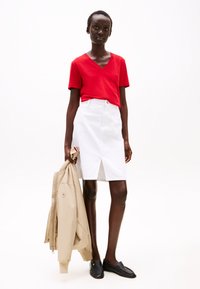 Raudonas V-kaklo marškinėliai, balta kelių ilgio sijonas su priekinėmis praplatėjimo detalėmis, smėlio spalvos bomberio striukė laikoma rankoje, juodi loaferiai, modelis su trumpais plaukais.