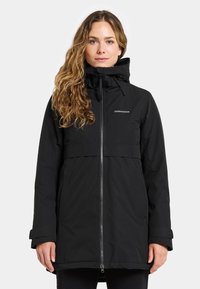 Woman with long wavy hair wearing a black Didriksons winter jacket with zipper and high collar, standing against a plain white background.