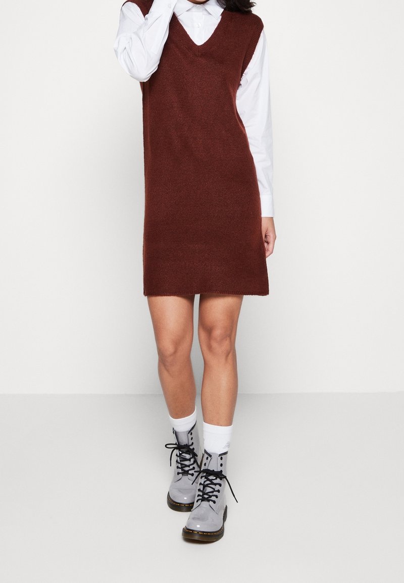 Personne portant une robe marron sans manches en tricot par-dessus une chemise blanche à manches longues, des chaussettes blanches et des bottines grises à lacets.