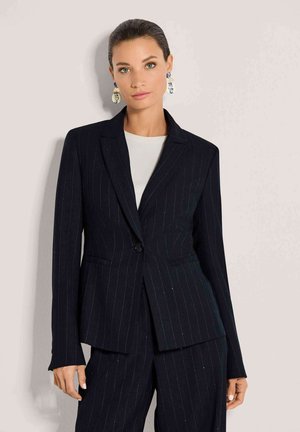 Femme aux cheveux tirés en arrière portant un costume noir à fines rayures et un haut blanc, debout devant un fond gris clair.