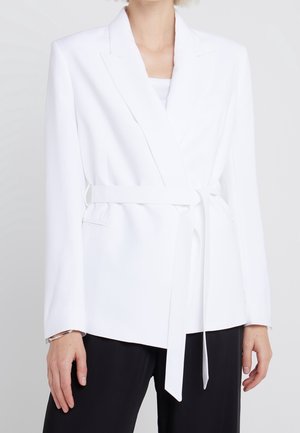 White belted blazer worn over a white top, paired with black pants, shown on a person from neck to hips against a plain background.