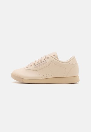 Baskets basses en cuir beige avec fermeture à lacets, perforations discrètes sur le côté et patch avec logo près des lacets, sur une semelle beige.