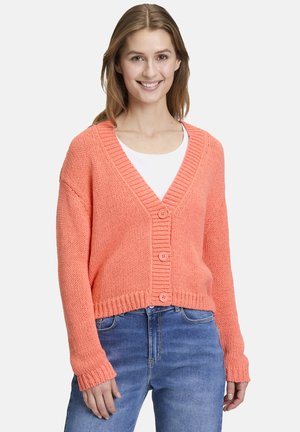 Korallenfarbener Strickcardigan mit tiefem V-Ausschnitt, fünf Knöpfen, gerippten Bündchen und verkürztem Saum. Über einem weißen Hemd und blauen Jeans getragen.