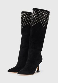 Botas negras de ante hasta la rodilla con puntera afilada, tacones delgados y adornos de tachuelas plateadas formando patrones diagonales cerca de la parte superior.