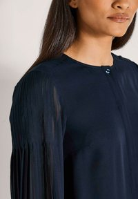 Navybluse mit rundem Ausschnitt, langen, gefältelten Netzärmeln und einem Knopfverschluss. Glatte Stoffoberfläche mit einem dezenten Glanz.
