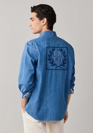 Man with dark hair wearing a blue denim shirt with embroidered laurel wreath and letters FA CO on the back, hands in pockets, light pants.