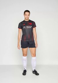 Čierny a červený futbalový dres s sieťovinovým dizajnom, logo na prednej strane, v kombinácii s čiernymi šortkami a bielymi ponožkami, dokončený čiernymi kopačkami.