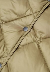 Tissu de veste beige matelassée avec un bouton-pression argenté visible et des coutures détaillées le long des coutures.