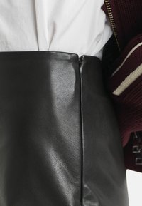 Zwarte leren rok met een gladde textuur, hoge taille en een rits aan de zijkant. Gecombineerd met een wit topje en een bourgondische cardigan.