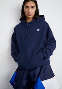 Ναυτικό μπλε βαμβακερή φόρμα με τσέπη μπροστά, κουκούλα με κορδόνι και άσπρο λογότυπο Nike στο στήθος. Διαθέτει χαλαρή εφαρμογή και ριπές στις μανσέτες.