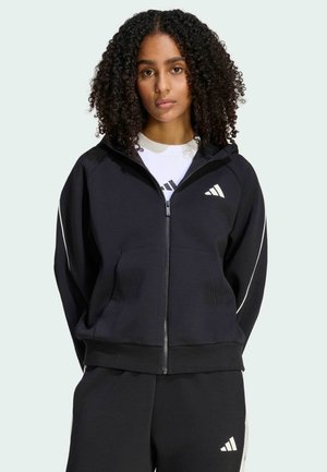 Ung kvinde med krøllet hår iført sort lynlåstrøje og matchende bukser med Adidas-logoer på bryst og ben.