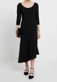 Svart knälång klänning med trekvartsärmar, rundad halsringning, figursydd bodice, utsvängd kjol och asymmetrisk fåll.