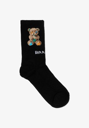 Chaussette noire arborant un ours en peluche brodé avec des patchs multicolores et un texte partiel blanc "BARB" près de la cheville.