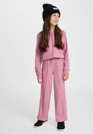Roze sweatshirt en broek set gemaakt van zachte stof, met een geweven jack, een elastische tailleband en zwarte sneakers. Een zwarte beanie maakt de look compleet.