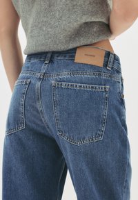 Osoba v modrých džínoch so zadnými vreckami a šedým úpletovým topom, ktorý je zasunutý, pričom je vidieť koženú značkovú nálepku na páse.